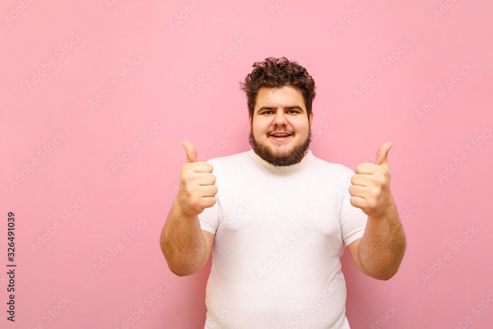 Happy young man in white t-shirt and overweight stands on a pink background and shows thumbs up with a smile on his face. Fat cheerful guy shows a Happy young man in white t-shirt and overweight stands on a pink background and shows thumbs up with a smile on his face. Fat cheerful guy shows a