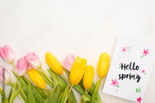 Top view of yellow, pink tulips and card with hello spring lettering on white background