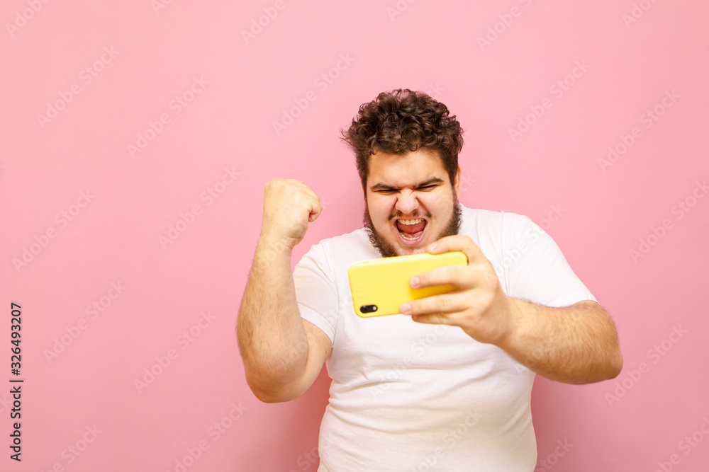 Portrait of winner gamer guy with smartphone in hands with raised hand rejoices in victory over pink background. Fat young man won a mobile game on a smartphone, happy looking in screen. Portrait of winner gamer guy with smartphone in hands with raised hand rejoices in victory over pink background. Fat young man won a mobile game on a smartphone, happy looking in screen.