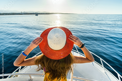 Young woman with long hair wearing yellow dress and straw hat standing on white yacht deck enjoying view of blue sea water.