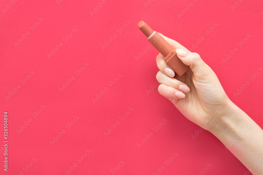 A girl's hand with a manicure holds a bright lipstick on a pink background. Red lipstick. Coral lipstick. Terracotta lipstick. Brick-colored lipstick. Makeup. Concept A girl's hand with a manicure holds a bright lipstick on a pink background. Red lipstick. Coral lipstick. Terracotta lipstick. Brick-colored lipstick. Makeup. Concept