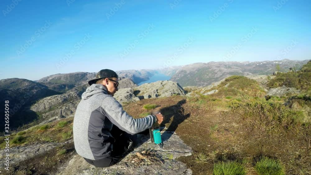 A young man cooking in the wilderness, nearby of Preikestolen, Norway, overlooking Lysefjord. Wild camping in the nature. The man cooks on a camp stove. He is enjoying peaceful and calm remedy. A young man cooking in the wilderness, nearby of Preikestolen, Norway, overlooking Lysefjord. Wild camping in the nature. The man cooks on a camp stove. He is enjoying peaceful and calm remedy.