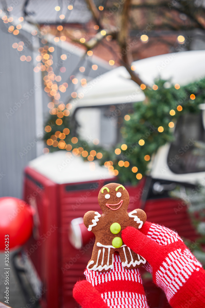 Hand in red mitten holding a smiling gingerbread man and christmas mood in blurred background. Christmas market in old town European small city.
