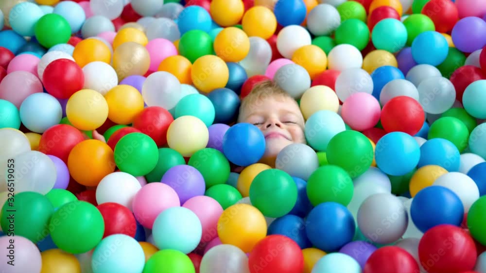 Blonde little boy lying on multi coloured plastic balls in big dry paddling pool in playing centre. Smiling at camera. Portrait close up. Having fun in playroom. Leisure Activity. Blonde little boy lying on multi coloured plastic balls in big dry paddling pool in playing centre. Smiling at camera. Portrait close up. Having fun in playroom. Leisure Activity.