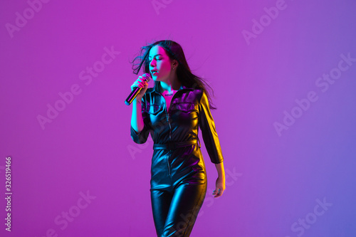 Caucasian female singer portrait isolated on purple studio background in neon light. Beautiful female model in black wear with microphone. Concept of human emotions, facial expression, ad, music, art.
