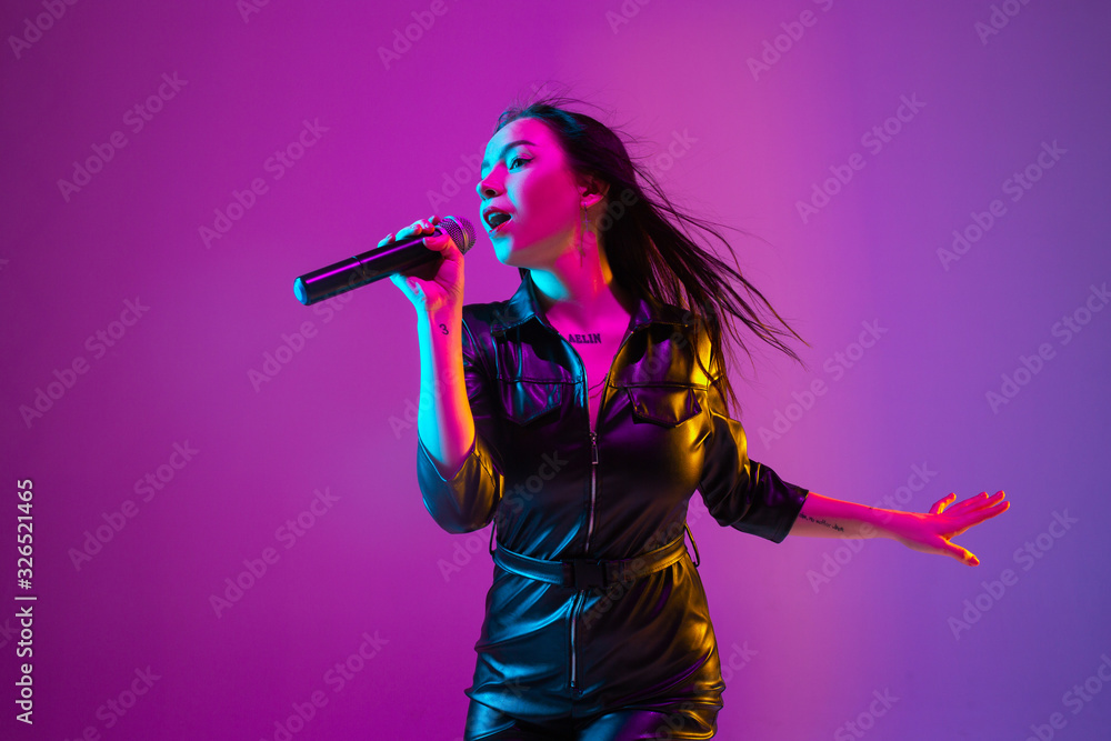 Caucasian female singer portrait isolated on purple studio background in neon light. Beautiful female model in black wear with microphone. Concept of human emotions, facial expression, ad, music, art. - obrazy, fototapety, plakaty 