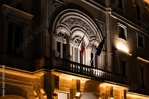 The facade of a Italian government building at night with the lit balcony displaying the Italian and European Union flags in Brindisi Italy.