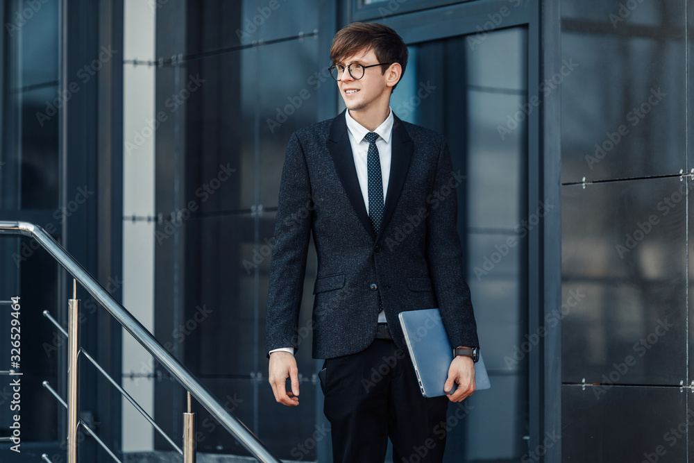 A handsome young businessman holds his laptop while standing in front of a glass building A handsome young businessman holds his laptop while standing in front of a glass building