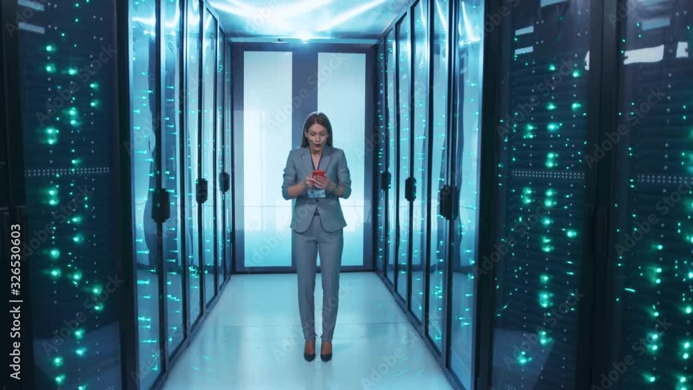 Attractive head IT administrator using a smartphone managing data center secure storage. In server room excited female worker having success reading mobile phone news. Attractive head IT administrator using a smartphone managing data center secure storage. In server room excited female worker having success reading mobile phone news.