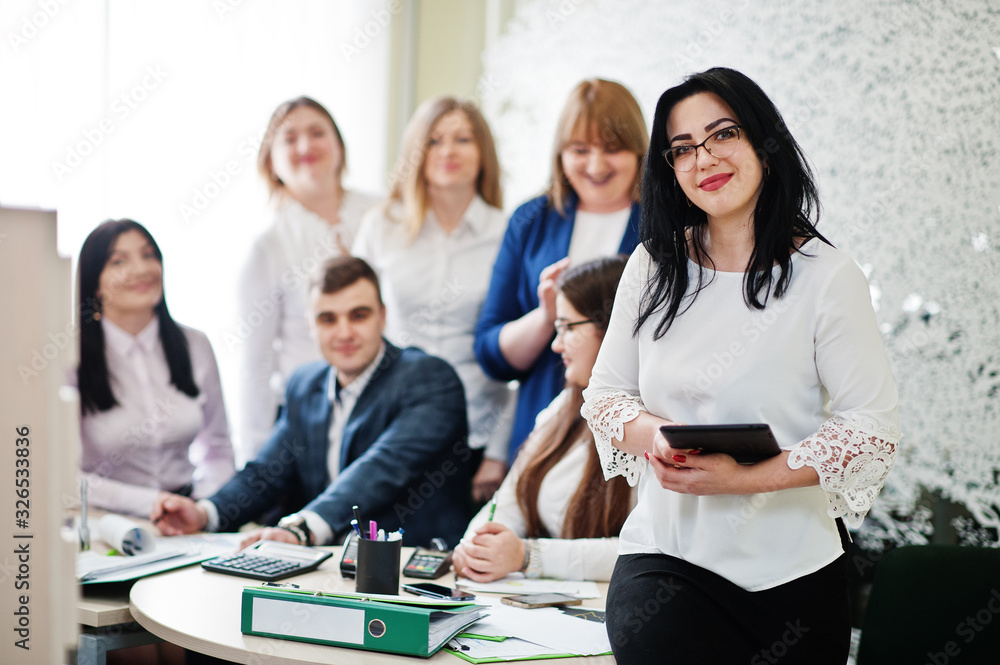Portrait of caucasian woman in white blouse and eyeglasses hold tablet against business people group of bank workers. Portrait of caucasian woman in white blouse and eyeglasses hold tablet against business people group of bank workers.
