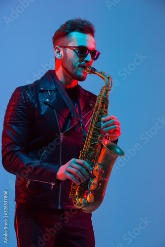 Photography Young caucasian jazz musician playing the saxophone on gradient blue-purple studio background in neon light