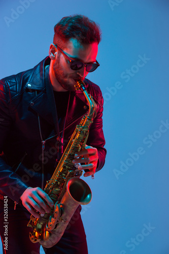 Young caucasian jazz musician playing the saxophone on gradient blue-purple studio background in neon light. Concept of music, hobby, festival. Joyful attractive guy. Colorful portrait of artist.