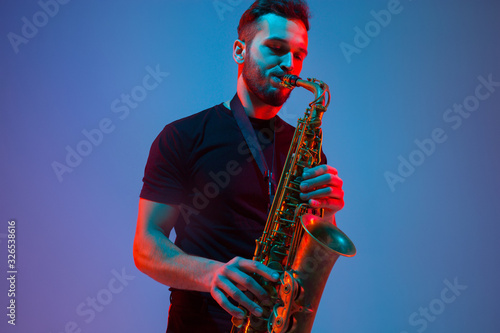 Canvas Print Young caucasian jazz musician playing the saxophone on gradient blue-purple studio background in neon light