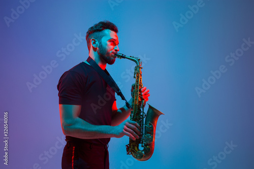 Young caucasian jazz musician playing the saxophone on gradient blue-purple studio background in neon light. Concept of music, hobby, festival. Joyful attractive guy. Colorful portrait of artist.
