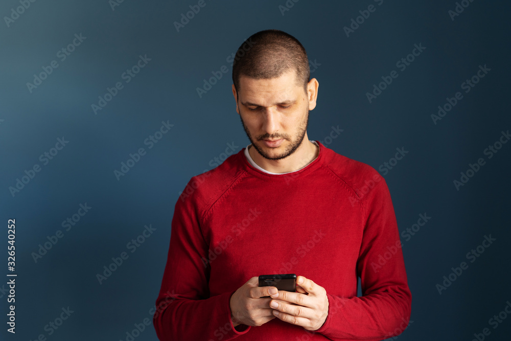 Caucasian man wearing red sweater standing in front of the blue background wall using smart phone mobile to send messages sms texting or browsing internet front view Caucasian man wearing red sweater standing in front of the blue background wall using smart phone mobile to send messages sms texting or browsing internet front view