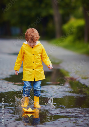 cute baby boy in yellow raincoat and rubber boots having fun, jumping in puddles, launching the paper boat after spring rain