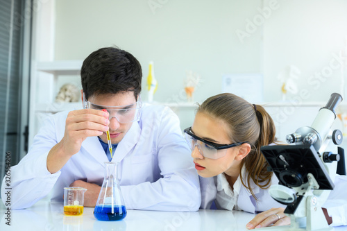 Young scientists run an experiment by dropping yellow liquid in blue liquid. Male and female scientists working together in laboratory room.