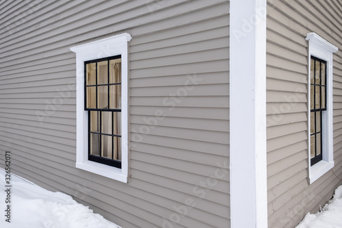 Two horizontal opening vintage windows in a tan colored wooden building. The windows have white trim around them. The exterior wall of the building is a beige clapboard. A snow drift is on the ground. photo