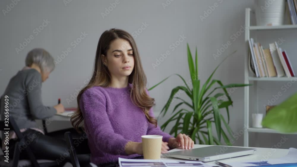 Panning left of tired thoughtful woman closing laptop and drinking takeaway coffee while working in office with colleague taking notes in background Panning left of tired thoughtful woman closing laptop and drinking takeaway coffee while working in office with colleague taking notes in background