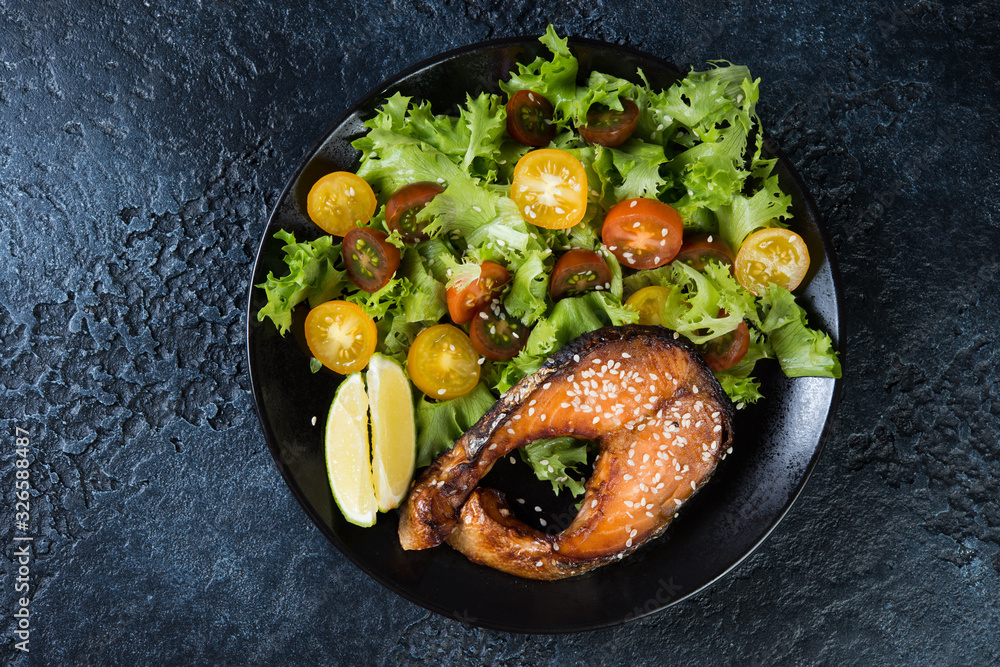 Salmon steak fried in teriyaki sauce with fresh lettuce and tomatoes on a black background. Top view, close-up Salmon steak fried in teriyaki sauce with fresh lettuce and tomatoes on a black background. Top view, close-up