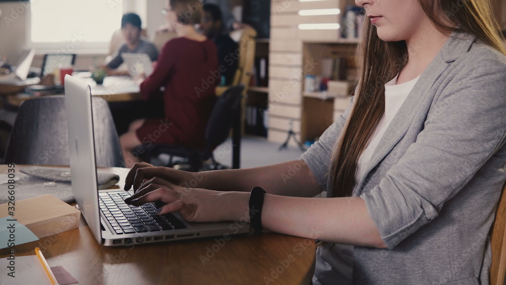 Concentrated European young girl typing a letter on laptop. Hard work in trendy office space, teamwork in background 4K.