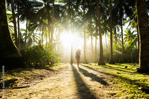 A beautiful  girl wearing a large hat and a red bikini is walking on a path surrounded by coconut palm trees illuminated by a stunning sunset in the background. Siargao Island, Philippines.
