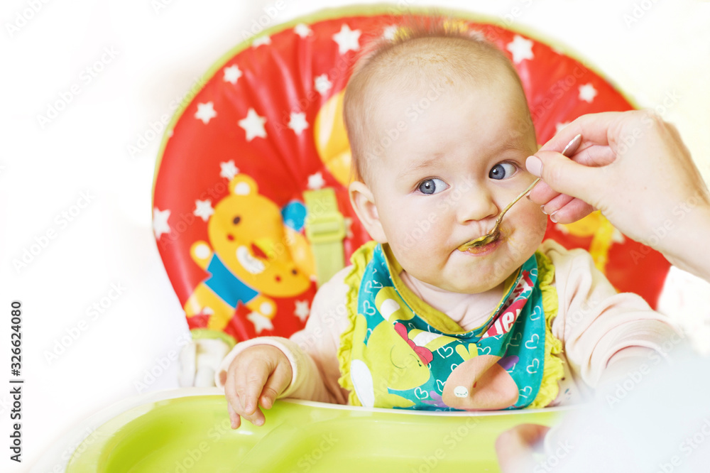 Mom feeds funny baby from a spoon on a white background. child eats in a highchair Mom feeds funny baby from a spoon on a white background. child eats in a highchair