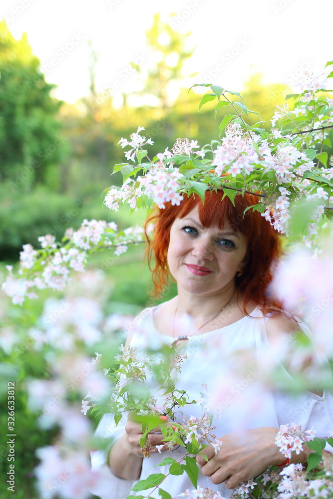 Naklejka premium beautiful red-haired Caucasian woman in a white dress enjoys flowering trees in the spring park 