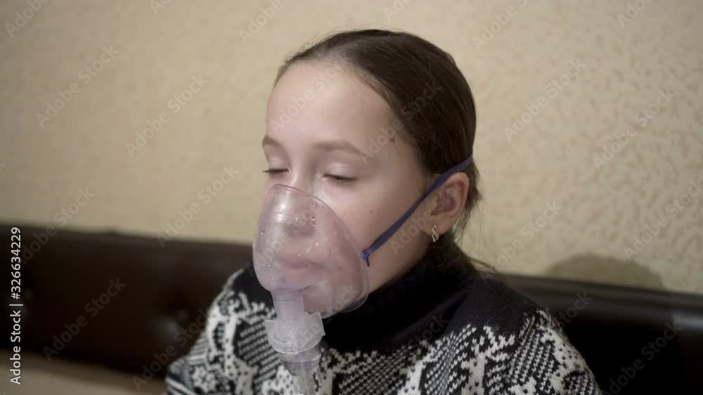 Sad little girl doing inhalation sitting at home at the table in a warm sweater. She adjusts the mask on her face and breathes through it. Close-up. 4K. Sad little girl doing inhalation sitting at home at the table in a warm sweater. She adjusts the mask on her face and breathes through it. Close-up. 4K.