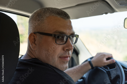 a man of 30-40 years old with a short haircut and gray hair with glasses sitting at the wheel of a car, looking at the camera