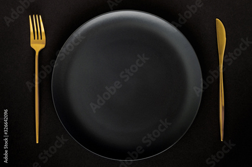 Black empty ceramic plate with golden cutlery (fork and knife) on a black background (table).