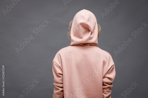A beautiful young girl stands with her back and turns around in a pink hoodie on a gray background.