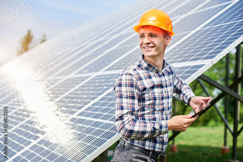 Happy guy in a helmet with a tablet in his hands looking away near the solar cell station with a reflection of the sun in them. Home construction. Green ecological power energy generation © anatoliy_gleb