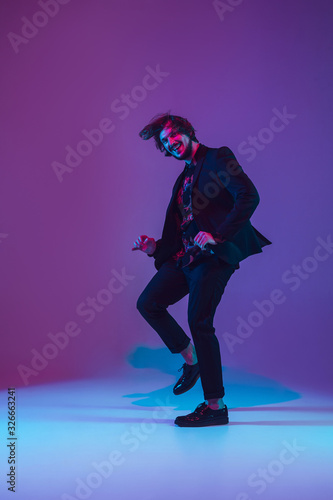 Young caucasian musician in casual dancing on gradient blue-purple background in neon light. Concept of music, hobby, festival. Joyful party host, DJ, stand upper, dancer. Colorful portrait of artist.