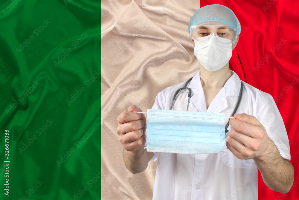 doctor in uniform with a stethoscope against the background of the silk national flag of italy holding a medical mask, concept of flu, virus, COVID-19, coronavirus, epidemic, vaccination doctor in uniform with a stethoscope against the background of the silk national flag of italy holding a medical mask, concept of flu, virus, COVID-19, coronavirus, epidemic, vaccination