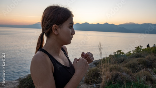 Young boxer girl posing by the sea over huge cliffs at sunset. She wears tights and sports bra.