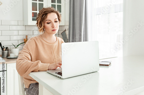 Cute woman using her notebook at the white table in bright kitchen