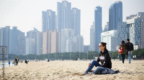 Young woman sits on the sandy beach in Busan city with skyscrapers of downtown on the background in South Korea, Asia.