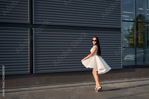 Portrait of beautiful fashionably dressed brunette woman wearing white stylish elegant dress and trendy sunglasses posing on grey wall background. Outdoors. Copy space.