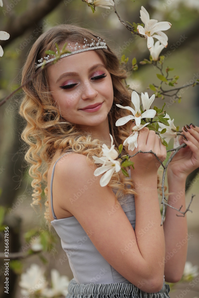 young beautiful blonde in a silver dress with a train in the spring garden, where white magnolia blooms, close-up young beautiful blonde in a silver dress with a train in the spring garden, where white magnolia blooms, close-up
