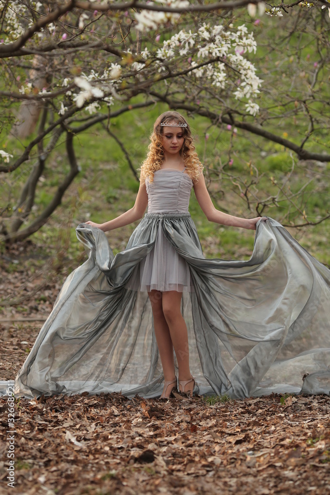  young beautiful blonde in a silver dress with a train in the spring garden, where white magnolia blooms
