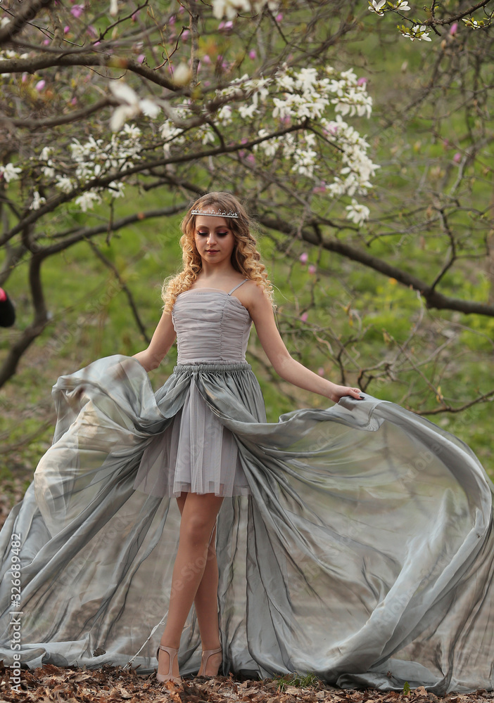 Fototapeta premium young beautiful blonde in a silver dress with a train in the spring garden, where white magnolia blooms