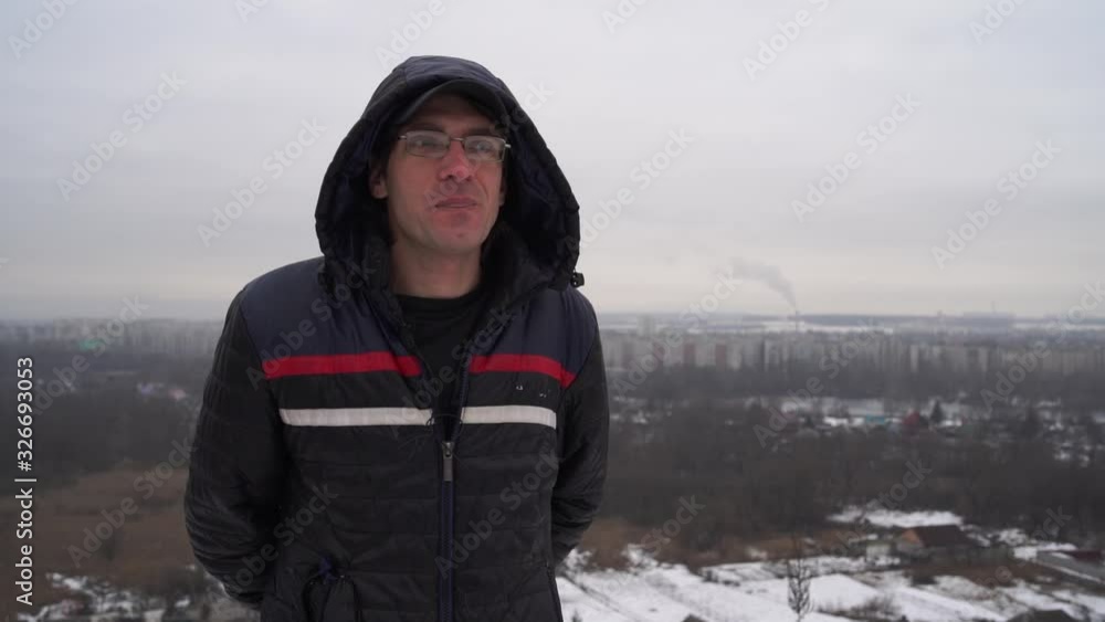 Mature man with glasses in hooded jacket looking thoughtfully into distance in cloudy weather. Adult male sniffing and thinking on background of panorama of small city. Mature man with glasses in hooded jacket looking thoughtfully into distance in cloudy weather. Adult male sniffing and thinking on background of panorama of small city.