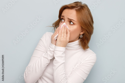 Studio portrait of scared young woman in white turtleneck with napkin blowing nose, looking to the source of allergy, place for advertising. Rhinitis, dust and pollen seasonal allergy. 