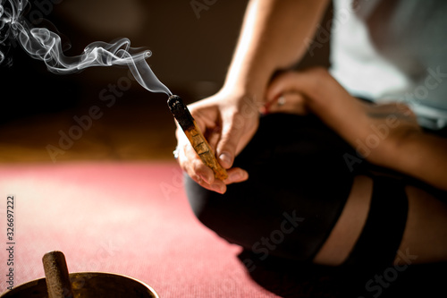 Woman in sport clothes practicing yoga in a studio room sitting in lotus pose torching the palo santo