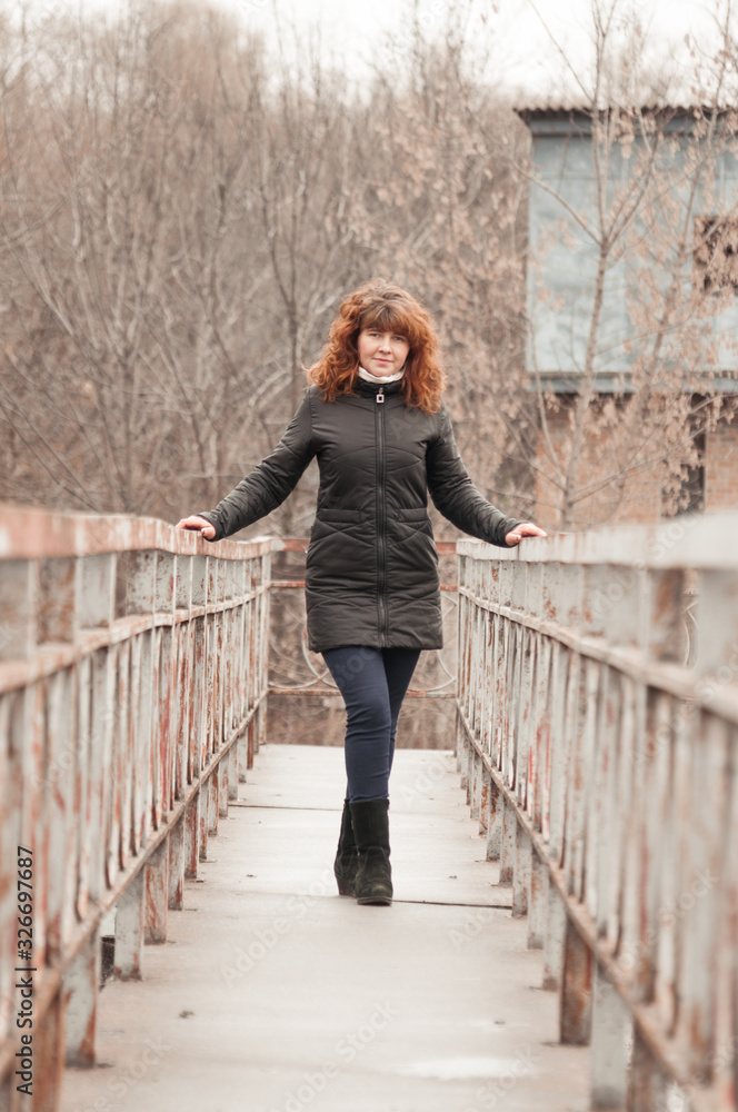 Young beautiful woman with red hair in a black jacket on a bridge in cloudy weather