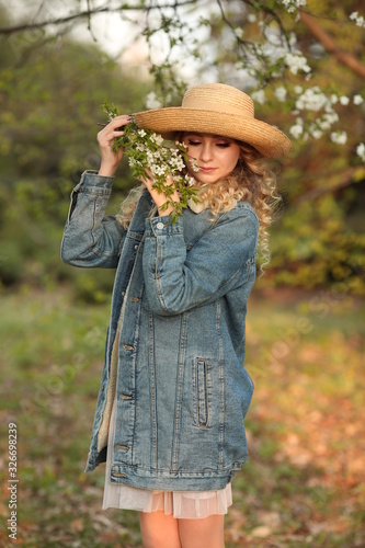  beautiful blonde girl with a bouquet of flowering branches in a straw hat and in a blue jeans jacket in a spring garden where trees bloom © Alina
