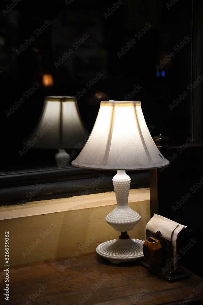 Table lamp. Elegant white antique table lamp closeup on dark blurry reflection. Lamp silhouette blurred reflection in glass window. Antique white lampshade glowing in darkness. Table lamp. Elegant white antique table lamp closeup on dark blurry reflection. Lamp silhouette blurred reflection in glass window. Antique white lampshade glowing in darkness.