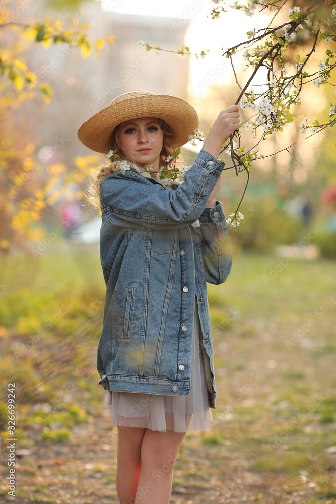  beautiful blonde girl with a bouquet of flowering branches in a straw hat and in a blue jeans jacket in a spring garden where trees bloom