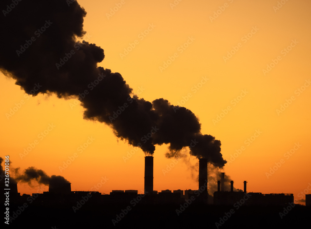 Silhouettes of the dark smoke clouds from tall chimneys of the industrial plant and clear sunset sky as background. Cityscape silhouette with twilight sky.